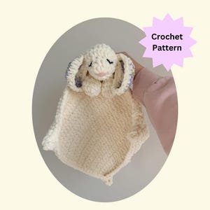 May include: A handmade crochet bunny lovey in cream and white. The bunny has long ears with lavender and yellow accents, closed eyes, and a pink mouth. The text "Crochet Pattern" is in a starburst shape.