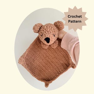 May include: A handmade crochet bear security blanket in a warm brown color. The bear has a black nose and small ears. The blanket is square with a textured surface and small corner accents. The text "Crochet Pattern" is visible.