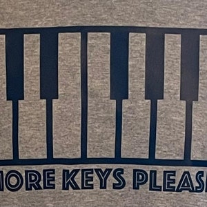 May include: A grey t-shirt with a black outline of a piano keyboard and the text "MORE KEYS PLEASE" printed on the front.