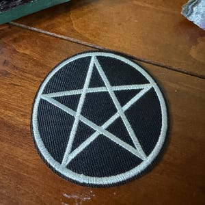 Peut inclure: Un écusson rond noir avec un pentagramme blanc. Le patch a une bordure cousue et est posé sur une surface en bois. Le pentagramme, une étoile à cinq branches, est un symbole souvent associé à l'occultisme.