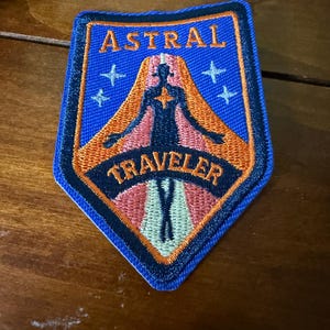 Op de afbeelding: Geborduurde patch met de woorden "ASTRAL TRAVELER" in zwarte letters. De patch heeft een blauwe achtergrond met een gestileerde figuur en oranje en roze accenten. Er zijn ook vier witte sterren aanwezig.