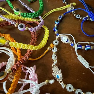 May include: A collection of colorful woven bracelets with various designs. The bracelets feature gold beads and protective eye charms in green, yellow, orange, purple, and white. Some bracelets have a Hamsa hand charm.