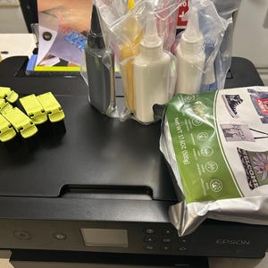May include: A black Epson printer with ink bottles and a paper package. The printer has a small display screen and control buttons. Ink bottles are in clear plastic packaging. A green paper package is also visible.