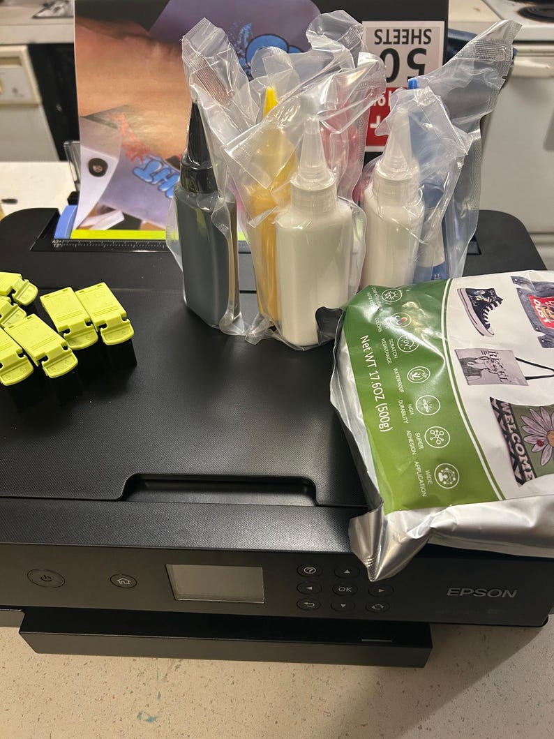 May include: A black Epson printer with ink bottles and a paper package. The printer has a small display screen and control buttons. Ink bottles are in clear plastic packaging. A green paper package is also visible.