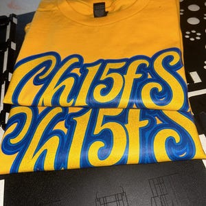May include: Folded yellow t-shirts with the word "Chiefs" in blue and white script. The shirts are stacked on top of each other, with the text prominently displayed. The shirts are on a black surface.