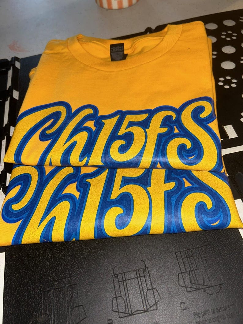 May include: Folded yellow t-shirts with the word "Chiefs" in blue and white script. The shirts are stacked on top of each other, with the text prominently displayed. The shirts are on a black surface.