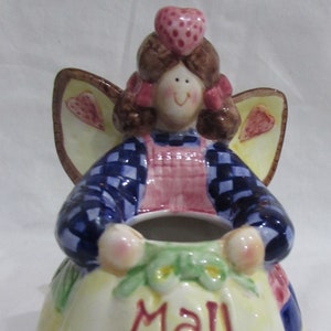 May include: A ceramic figurine of an angel with wings, wearing a blue and white checkered dress, a pink apron, and a red heart on her head. The angel is holding a yellow container with the words "Mail Money" written on it.