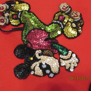 May include: A colorful sequined appliqué of a cartoon character, possibly Mickey Mouse, with black, green, gold, pink, and white sequins. The appliqué is pinned to a red fabric background.