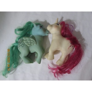 May include: Two vintage toy unicorns. One is light green with green hair and wings, the other is off-white with pink hair. The green unicorn has musical notes on its wing. Both have a horn and are facing each other.