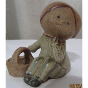 Może przedstawiać: Ceramiczna figurka siedzącego dziecka z koszem. Figurka ma jasnobrązową głowę i zielono-brązowy strój. Dziecko trzyma się za podbródek, a kosz jest obok niego.