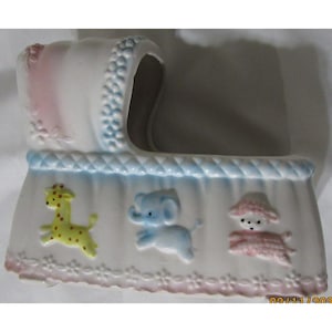 Puede incluir: Una maceta de cerámica con forma de cuna de bebé en blanco, rosa y azul. La cuna presenta imágenes en relieve de una jirafa, un elefante y un bebé con gorro. La maceta tiene un borde decorativo y está diseñada para decoración infantil.