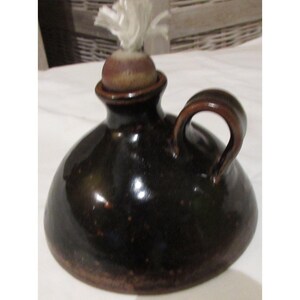 May include: A dark brown ceramic oil lamp with a handle. The lamp has a round body and a small opening at the top with a cork and a white wick. The lamp is glazed and has a glossy finish.