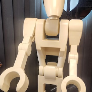 以下が含まれることがあります： 人型デザインの、オフホワイトのレゴ風ロボットの大きなフィギュア。長方形の胴体、爪のような手を持つ関節式アーム、丸みを帯びた頭部が特徴です。背景は、白と黒のデザインの濃い灰色の壁です。