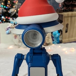 May include: A blue robot figure wearing a red and white Santa hat. The robot has a cylindrical head with a large eye-like lens. The background includes a Christmas tree and a snowy landscape, suggesting a holiday theme.