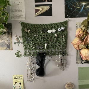 May include: A green crocheted wall hanging with a variety of jewelry and accessories hanging from it. The hanging is attached to a white wall with a white and green floral sticker. There are also several other decorations on the wall, including a poem, a picture of Saturn, and a picture of the solar system.