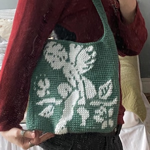 Op de afbeelding: Een groene gehaakte tote bag met een wit bloemenontwerp. De tas heeft een schouderband en is gemaakt van een getextureerd garen. Het ontwerp heeft een cherubijn en bloemelementen. De tas wordt door een persoon vastgehouden.
