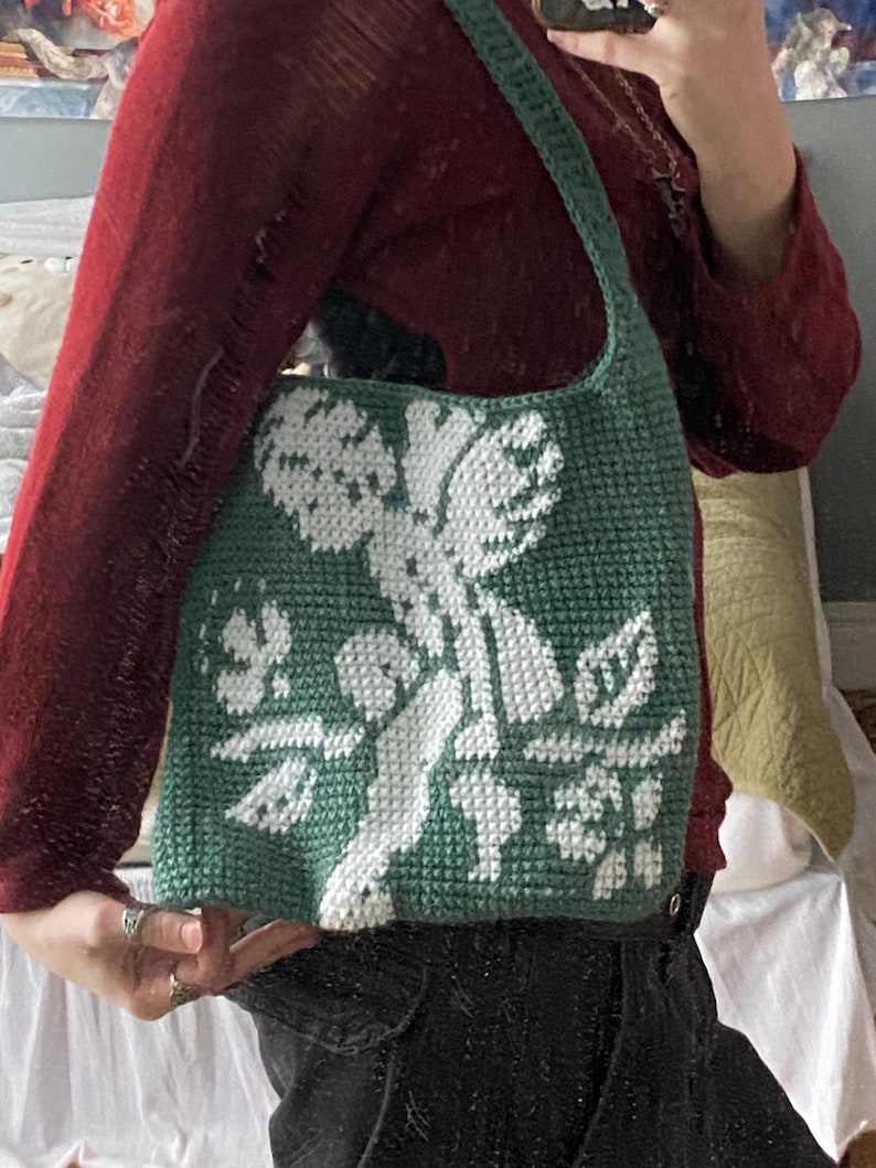 Op de afbeelding: Een groene gehaakte tote bag met een wit bloemenontwerp. De tas heeft een schouderband en is gemaakt van een getextureerd garen. Het ontwerp heeft een cherubijn en bloemelementen. De tas wordt door een persoon vastgehouden.
