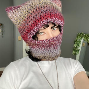 May include: A red, purple, and yellow crocheted cat ear beanie with a matching face mask. The beanie is made with a soft, fuzzy yarn.