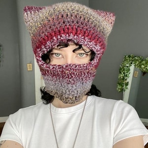 May include: A hand-crocheted balaclava featuring pointed cat-ear details at the top. The yarn is a variegated blend of deep burgundy, muted purple, and earthy brown tones, worked in a textured stitch pattern that covers the head and neck area.