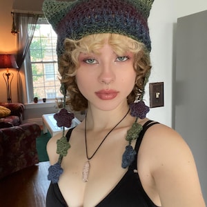 Puede incluir: Una mujer con un gorro de punto de ganchillo de color verde, morado y azul con orejas de gato y adornos florales. También lleva un top negro sin mangas y un collar con un colgante de cristal.