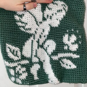 Op de afbeelding: Een handgemaakte, groene gehaakte tas met een wit bloemenontwerp. De tas heeft een gedetailleerd, gestructureerd patroon van bloemen en bladeren, dat de kunst van het haken laat zien. Het kleurenpalet van de tas is een opvallend contrast van groen en wit.