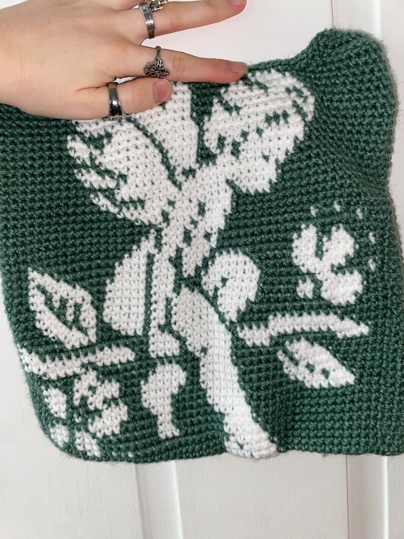 Op de afbeelding: Een handgemaakte, groene gehaakte tas met een wit bloemenontwerp. De tas heeft een gedetailleerd, gestructureerd patroon van bloemen en bladeren, dat de kunst van het haken laat zien. Het kleurenpalet van de tas is een opvallend contrast van groen en wit.