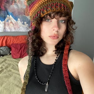 May include: A handmade crochet hat with cat ears, featuring a blend of warm colors like orange, brown, and green. The hat has long, braided ties that cascade down the sides. The hat is worn with a black tank top and a crystal pendant necklace.