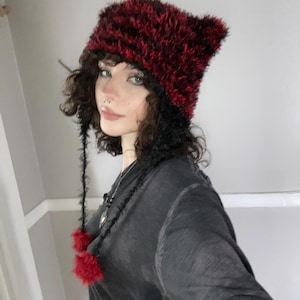 May include: A red and black faux fur hat with ear-like points. The hat has long black ties with red pom-poms. The hat is worn with a black long-sleeved shirt.