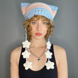 Puede incluir: Un gorro de crochet hecho a mano con orejas de gato en azul pastel, rosa y blanco. El gorro tiene cuerdas azules colgantes con detalles de estrellas blancas. La persona lleva una camiseta sin mangas negra y un collar plateado con un colgante de corazón.