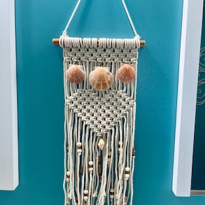 May include: A macrame wall hanging with a wooden dowel at the top. The hanging features a triangular pattern with white rope and three seashells. The bottom of the hanging has white and brown beads.