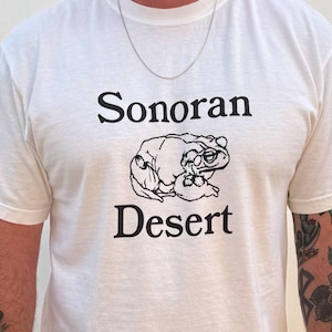 May include: White t-shirt with the words "Sonoran Desert" in black, above and below a black and white illustration of a frog wearing glasses. The shirt is short-sleeved.