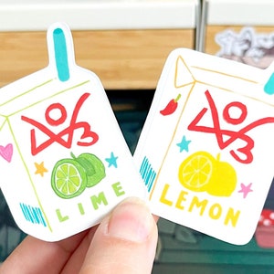 May include: Two white stickers shaped like juice boxes, one with a lime and the word "LIME", the other with a lemon and the word "LEMON". Both have a red design, blue straw, and colorful accents.