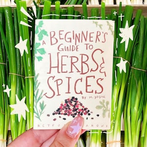 May include: A hand holds a book titled "A Beginner's Guide to Herbs & Spices" with a colorful illustration of spices. The book is surrounded by fresh green onions tied in bundles. The text is in a handwritten style.