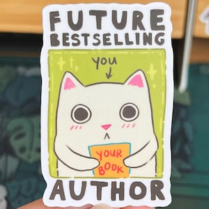 Könnte beinhalten: Ein weißer Aufkleber mit grünem Hintergrund, auf dem eine Comic-Katze zu sehen ist, die ein Buch mit der Aufschrift "Your Book" hält. Der Text auf dem Aufkleber lautet "Future Bestselling Author" mit einem Pfeil, der nach unten auf die Katze und das Wort "You" zeigt.