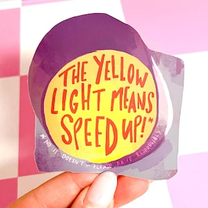 May include: A sticker with a purple border and a yellow circle in the center. The text "THE YELLOW LIGHT MEANS SPEED UP!" is written in red. The sticker also includes the text "* NO IT DOESN'T - PLEASE DRIVE RESPONSIBLY."
