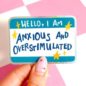 Könnte beinhalten: Ein rechteckiger Aufkleber mit türkisblauem Hintergrund und silbernem Glitzerbesatz. Der Aufkleber trägt den Text "HELLO, I AM ANXIOUS AND OVERSTIMULATED" in blauer und gelber Schrift. Der Aufkleber wird von einer Hand mit rosa Nagellack gehalten.
