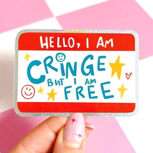 Puede incluir: Una pegatina rectangular con la parte superior roja y un borde plateado brillante. La pegatina dice "HELLO, I AM CRINGE BUT I AM FREE" en letras blancas y turquesas, con estrellas amarillas y una carita sonriente.