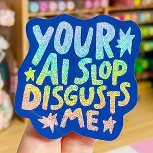 May include: A die-cut sticker with a deep blue background featuring the holographic text "YOUR AI SLOP DISGUSTS ME" in a bold, hand-lettered style. The design includes small holographic star accents scattered around the text.