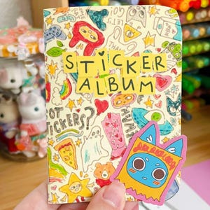 Könnte beinhalten: Ein kleines Sticker-Album mit cremefarbenem Einband, verziert mit handgezeichneten Motiven wie Geistern, Sternen, Pizza und Regenbögen. Der Text STICKER ALBUM steht auf gelben Papierstreifen, ergänzt durch einen blauen Katzen-Sticker.