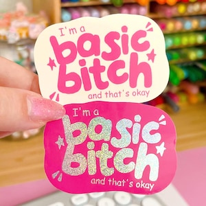 Op de afbeelding: Twee gestanste stickers met afgeronde randen. De bovenste sticker is crèmekleurig met felroze tekst die I'm a basic bitch and that's okay leest. De onderste sticker is felroze met holografische zilveren tekst die I'm a basic bitch and that's okay leest.