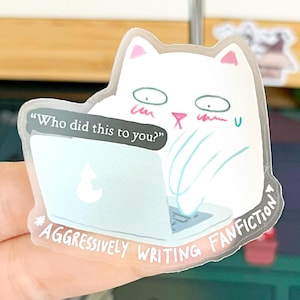 May include: A clear sticker featuring a white cat with pink ears and a surprised expression, typing on a laptop. Text on the sticker reads "Who did this to you?" and "Aggressively Writing Fanfiction."