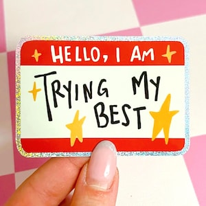 Puede incluir: Una pegatina rectangular con borde rojo y centro blanco. La pegatina dice "HELLO, I AM" en blanco y "TRYING MY BEST" en negro. Estrellas y detalles dorados decoran la pegatina. La pegatina tiene un borde brillante e iridiscente.