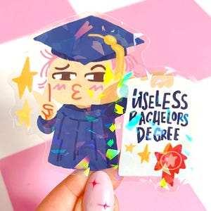 Op de afbeelding: Een holografische sticker met een stripfiguur-afgestudeerde in een blauwe baret en toga, met een diploma met de tekst "USELESS BACHELORS DEGREE". De afgestudeerde geeft een vredesteken en een kusje. Gouden sterren zijn verspreid.