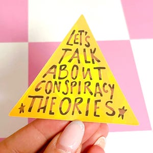 May include: A yellow triangular sticker with the text "LET'S TALK ABOUT CONSPIRACY THEORIES" in black and brown lettering. The sticker has a star on each side of the text. The background is a pink and white checkered pattern.