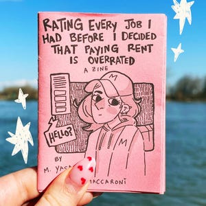 Op de afbeelding: Een roze zine met de titel "RATING EVERY JOB I HAD BEFORE I DECIDED THAT PAYING RENT IS OVERRATED". De cover toont een zwart-wit illustratie van een persoon met een hoed. De zine wordt vastgehouden tegen een achtergrond van blauwe lucht en water.
