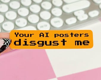 Puede incluir: Una pegatina rectangular naranja con esquinas redondeadas que presenta un texto negro pixelado que dice Your AI posters disgust me. La pegatina tiene un acabado mate suave y se sostiene frente a un fondo borroso de un teclado blanco.