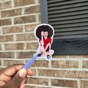 May include: A sticker featuring a cartoon illustration of a woman with an afro hairstyle wearing a red shirt and sitting with her legs crossed. She is wearing glasses and has a relaxed expression.