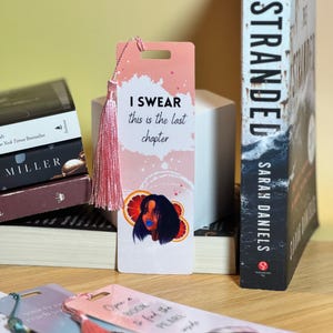 May include: A bookmark with a pink tassel and the text "I SWEAR this is the last chapter." The bookmark features an illustration of a woman with blue lips and orange slices. A stack of books and a book titled "STRANDED" are in the background.