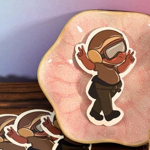 Puede incluir: Un personaje de dibujos animados marrón y blanco que lleva un casco marrón y gafas de protección, con un signo de paz en cada mano, se muestra en un plato de cerámica rosa y blanco.