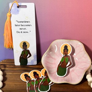 May include: A bookmark with a quote that reads "Sometimes later becomes never. Do it now." and a colorful illustration of a woman with green leaves. The bookmark is on a wooden surface with a pink dish and stickers of the same illustration.